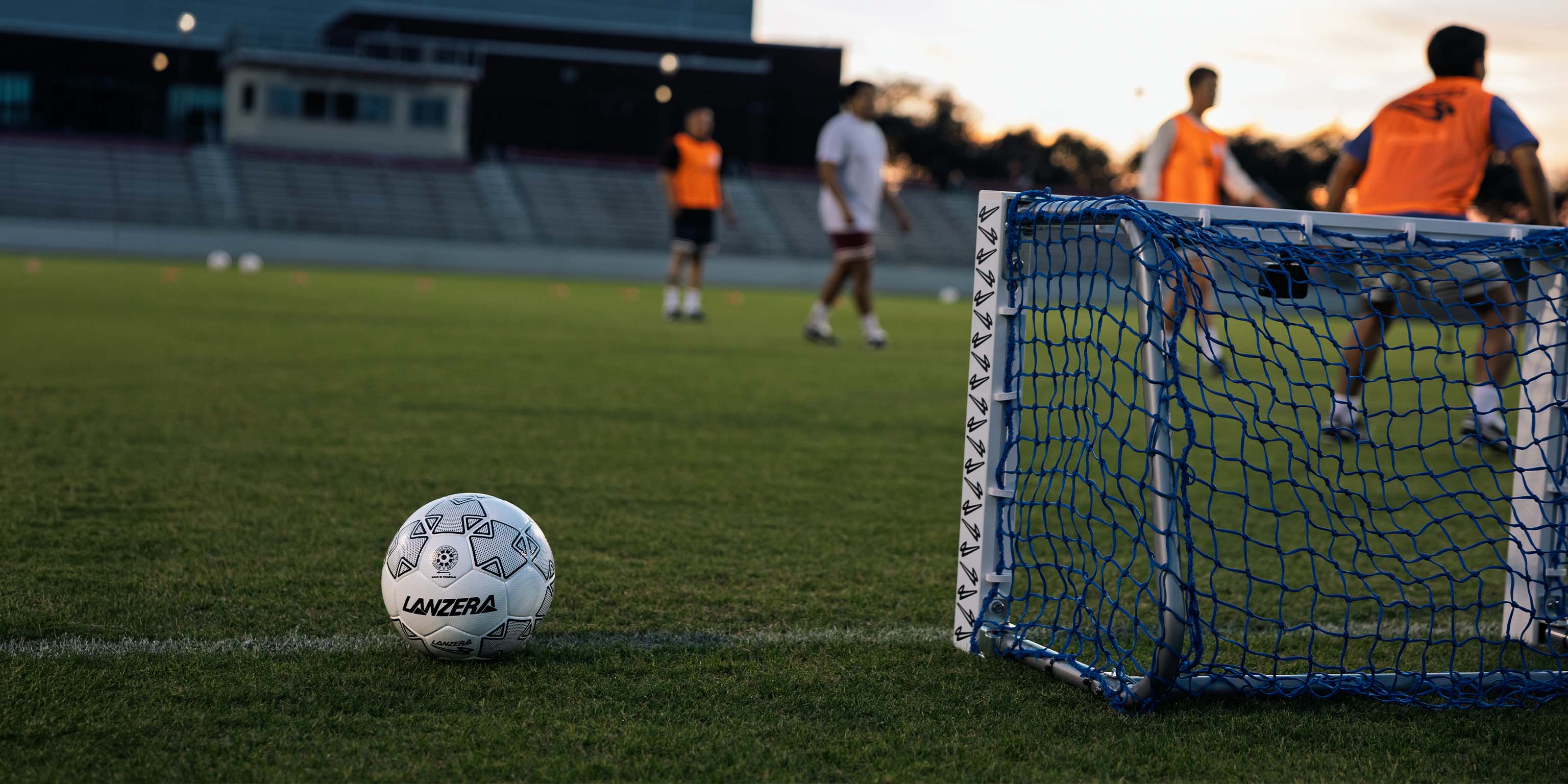 A Lanzera Campione 30 soccer ball with the logo visible on its side sits on a green grass field in the foreground, with a blue soccer goal on the right and players in orange jerseys blurred in the background during a sunset.