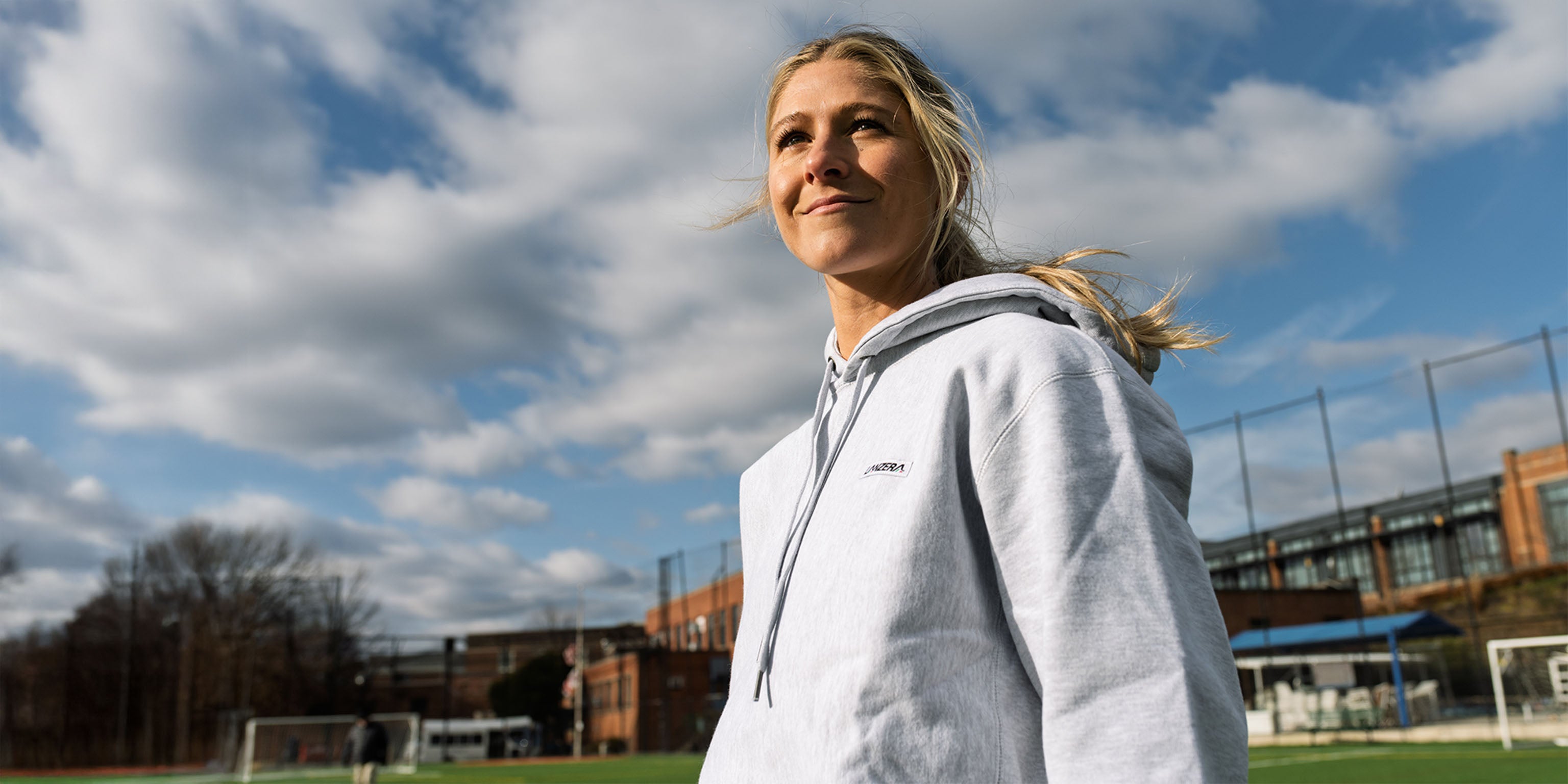 Woman wearing the Lanzera Club Hoodie in grey at a football pitch on a clear day, representing Lanzera’s everyday football lifestyle.