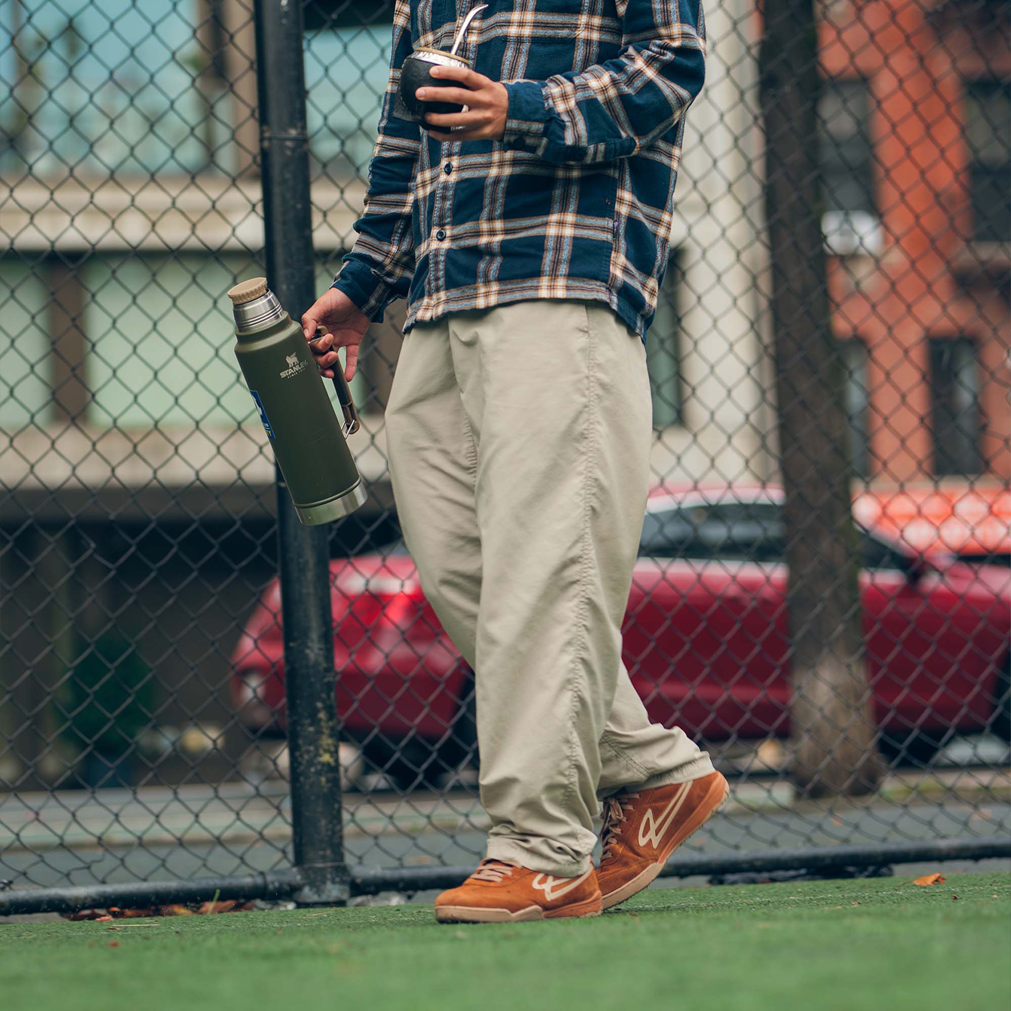 Person holding a thermos and mate, walking on a soccer field wearing Lanzera Jackal sneakers in caramello brown with a chain-link fence in the background.