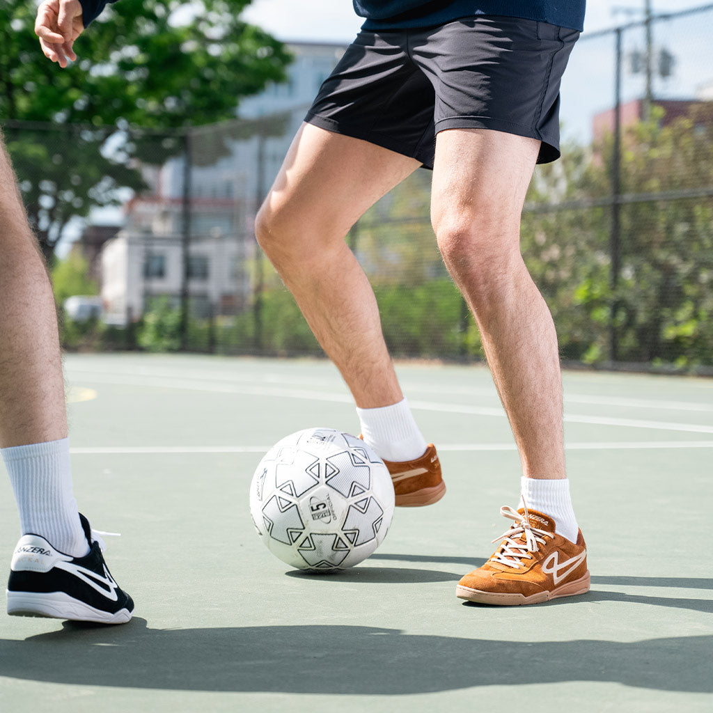 Two players competing on a court in Lanzera Jackal sneakers, emphasizing the futsal-ready traction, responsive fit, and streetwear-inspired design of the Caramello brown colorway.