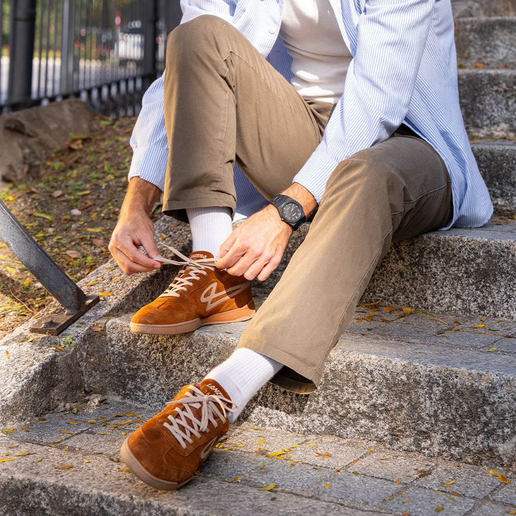 A person tying the laces of the Lanzera Jackal Caramello sneakers on outdoor stone steps, emphasizing the timeless suede design and versatile styling for streetwear and soccer culture.