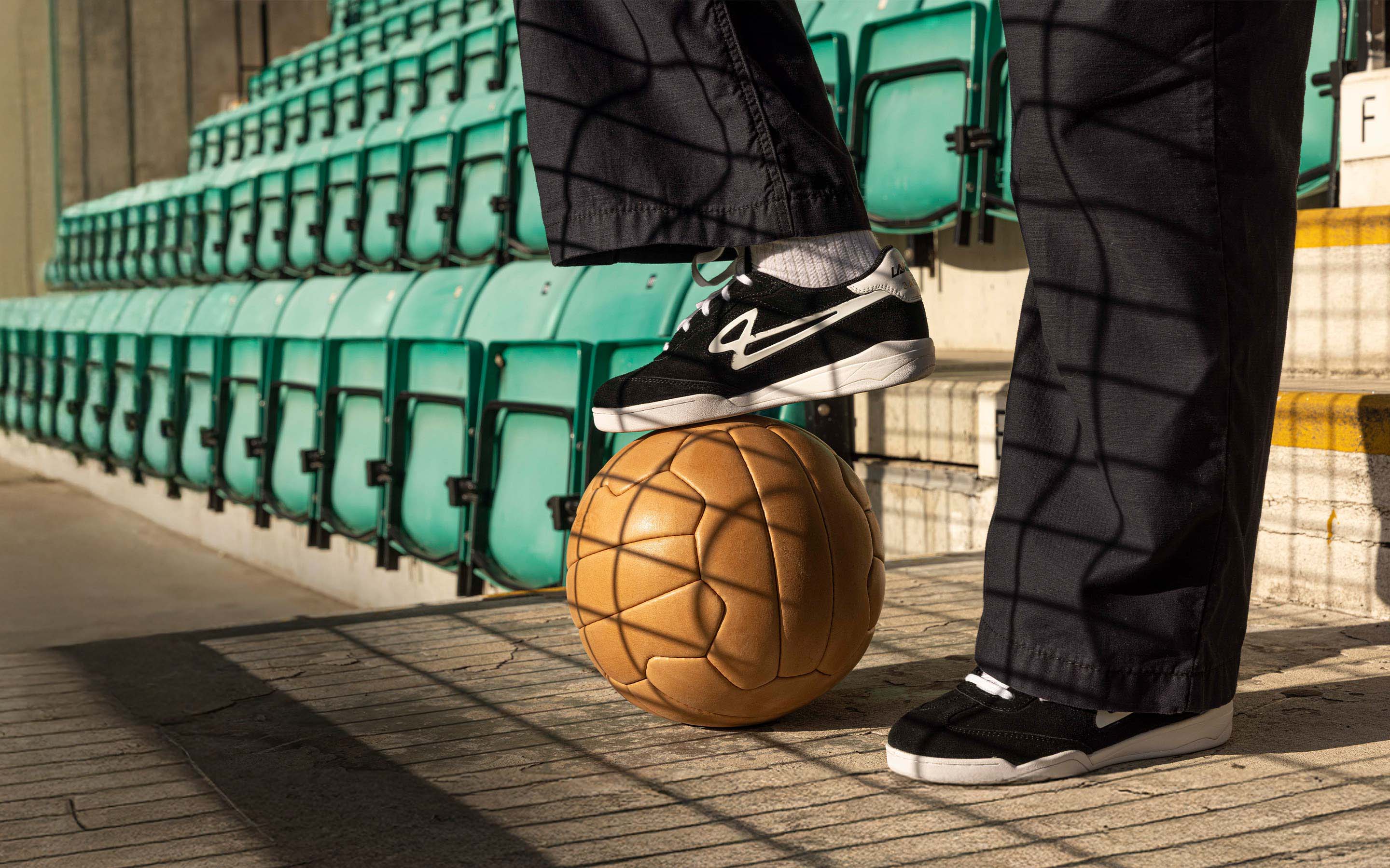 A close up of a person wearing Lanzera Jackal suede leather sneakers at a stadium, photographed with golden light and a classic football.
