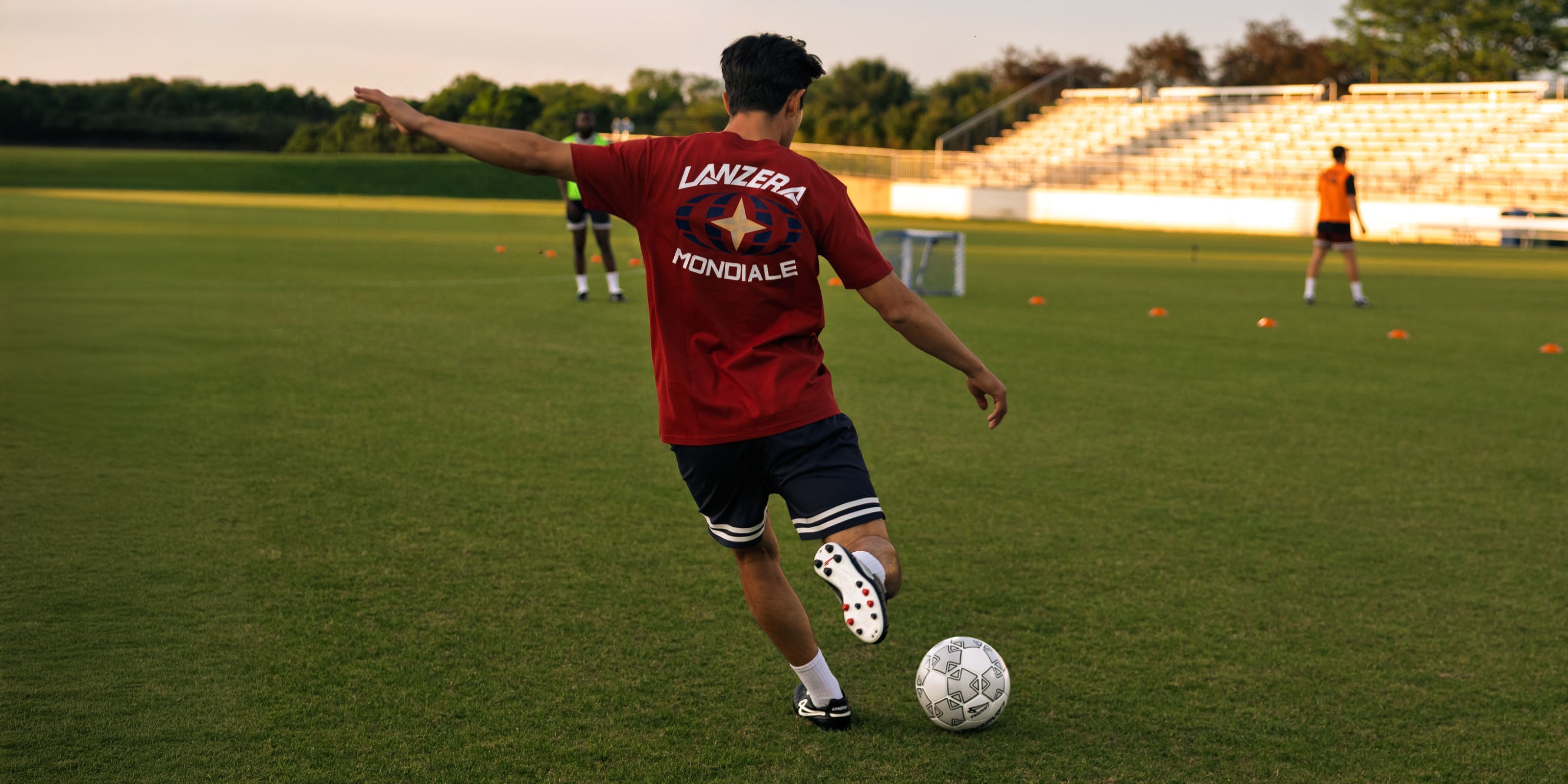 Player in a red Lanzera Mondiale Tee kicking a Lanzera soccer ball on a field with other players in the background.