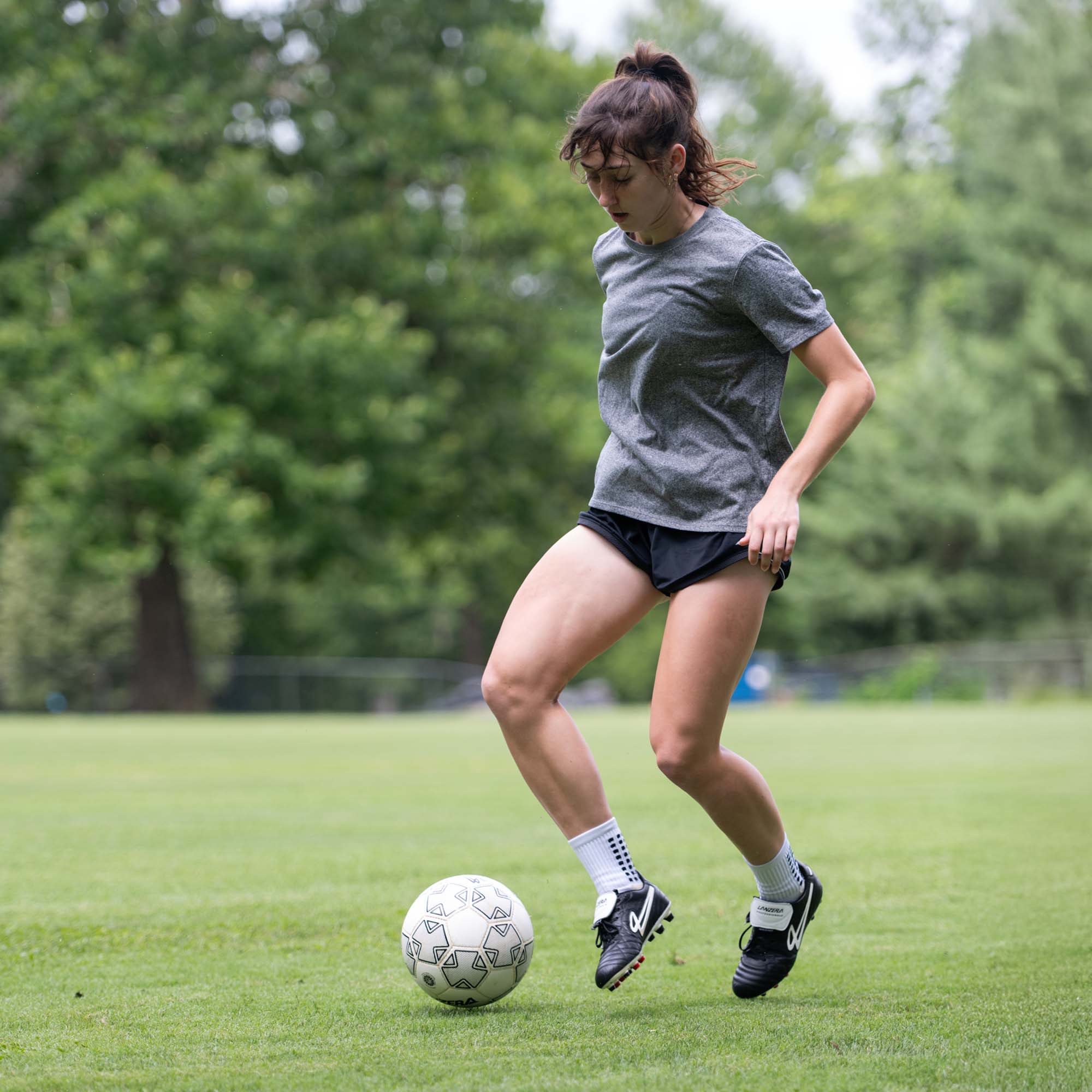 Footballer wearing Lanzera Professionale cleats, dribbling on a grass field with trees in the background.