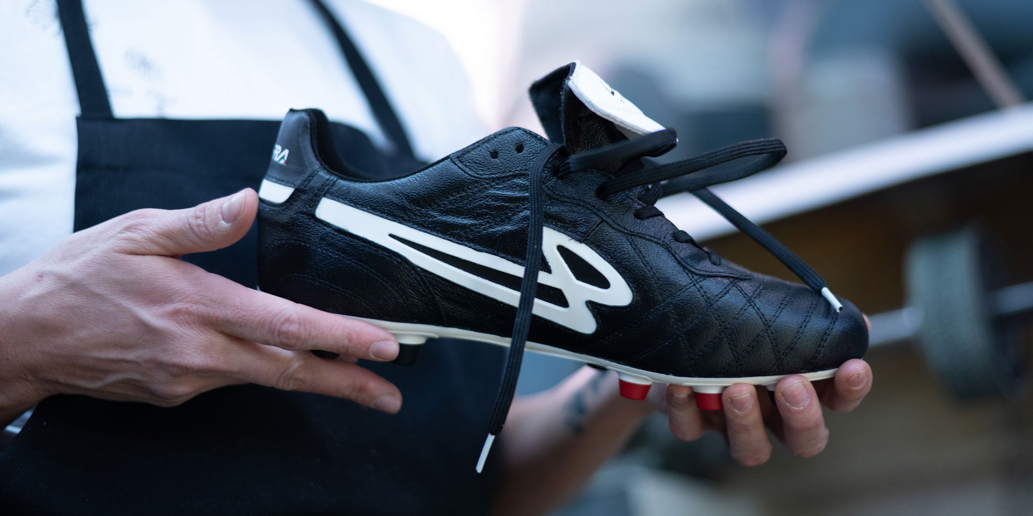 A close-up of the Lanzera Professionale soccer cleat, handcrafted in Italy from premium K-leather. A skilled artisan holds the boot, showcasing its meticulous stitching, iconic fold-over tongue, and signature Lanzera branding. This image represents the craftsmanship and heritage behind every pair of Professionale cleats.