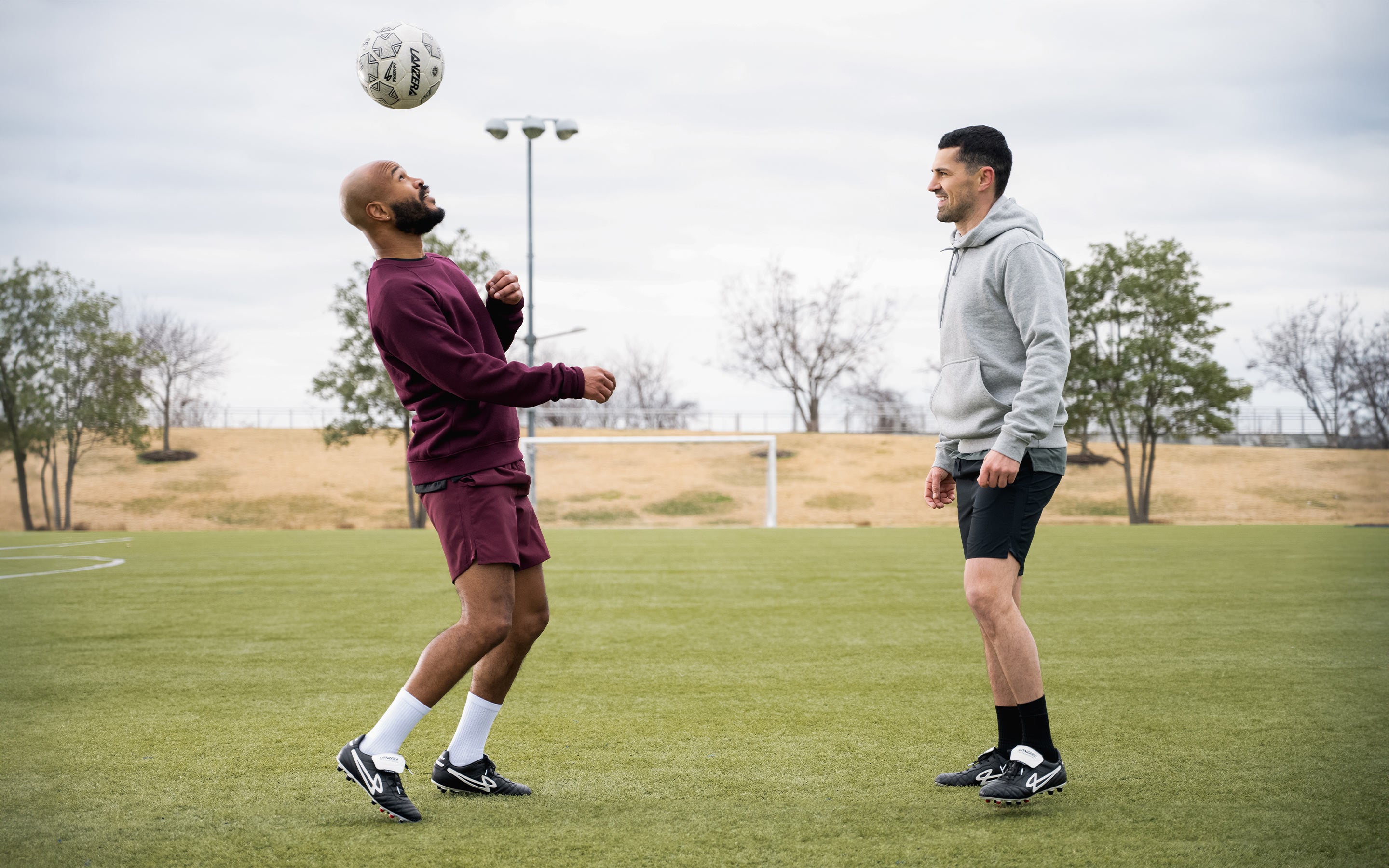 Two footballers on the pitch wearing Lanzera Professionale black K-leather soccer cleats, as one performs a header. Designed for precision, comfort, and firm ground performance, these boots blend heritage craftsmanship with modern football culture.