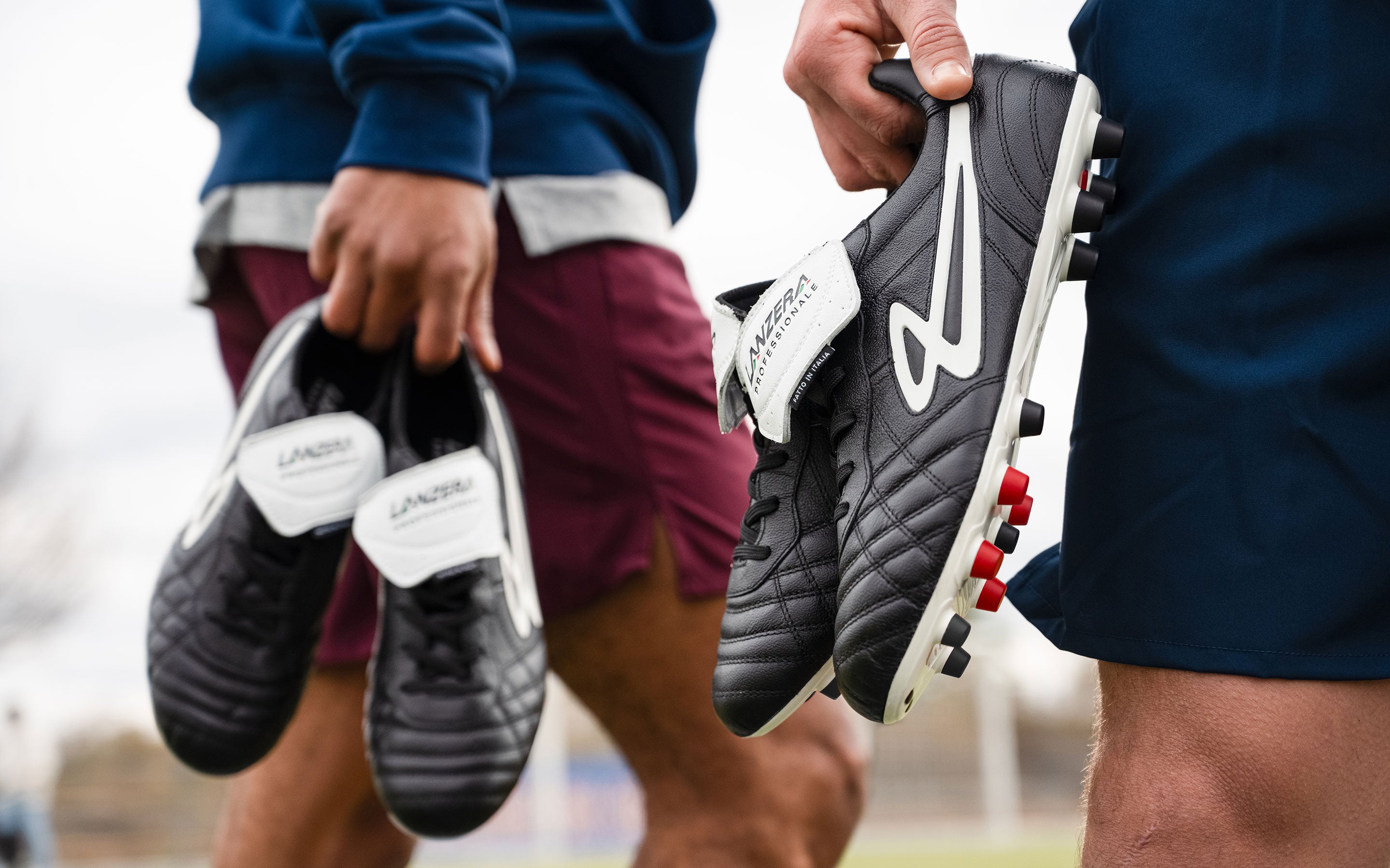 Two players holding Lanzera Professionale black K-leather soccer cleats, featuring a premium leather upper, fold-over tongue, and firm ground soleplate designed for ultimate control and precision on the pitch.