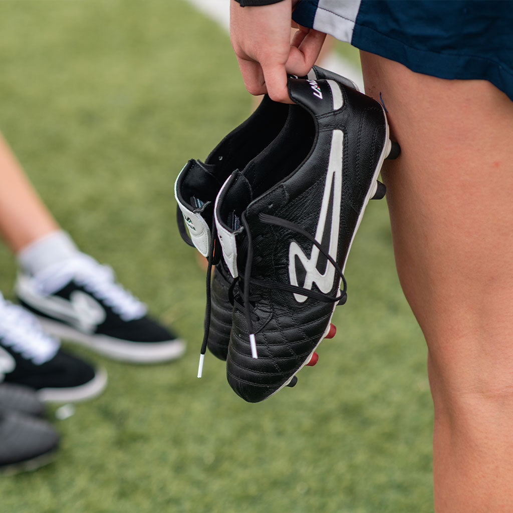 A player holding the Lanzera Professionale black K-leather soccer cleats, showcasing the premium kangaroo leather upper, fold-over tongue, and quilted stitching for superior touch and control.